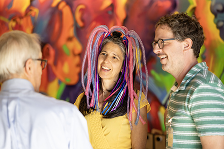 Woman with colorful light beads hanging from hair talking to two men in front of colorful wall mural.